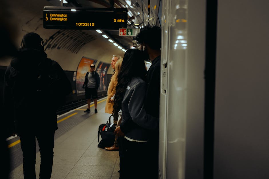 Photograph of a busy underground train station platform showing several passengers waiting for their train. Some individuals are wearing backpacks and casual clothing, with one woman holding a handbag, all standing near the edge of the platform. The station features tiled flooring, a safety yellow line, and overhead digital signage displaying train destinations and times, including Kennington with an estimated arrival in five minutes. The scene is lit by artificial lighting, and a train with open doors is visible in the background. This setting illustrates typical urban transportation infrastructure, relevant to home relocation and moving logistics services offered by Man and a Van Kennington, emphasizing the importance of efficient travel arrangements during house removals and furniture transport.