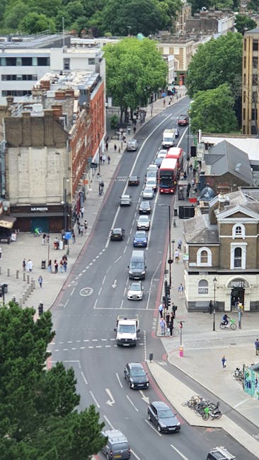 An aerial view of a busy residential street in Kennington with a row of parked cars along the curb and a line of moving vehicles on the road, including a red double-decker bus. The street is lined with trees and a mix of Victorian and modern buildings, some with flat roofs and others with pitched roofs. Pedestrians are walking on the sidewalks, and bicycles are parked near a safe crossing area close to a small, white, Victorian-style detached house. The scene captures the typical urban environment associated with house removals and furniture transport services, with a clear view of the road, parked and moving vehicles, street infrastructure, and nearby residential properties supporting effective home relocation logistics. Such detailed visual contexts assist in understanding vehicle loading, packing, and moving processes, relevant to services offered by Man and a Van Kennington, especially in busy city settings.