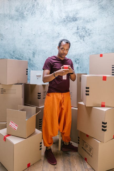 A man wearing an orange pair of trousers and a maroon t-shirt stands amidst several cardboard boxes, some of which are open and partially filled, in a room with a textured light blue wall. He is holding a smartphone in both hands, appearing to be checking information or coordinating a move. The boxes are of various sizes, and some have red and black packing labels or tape affixed. The scene suggests a home relocation or packing process, with the man possibly preparing for or managing a furniture transport or moving service. The environment is well-lit, and the setting indicates an indoor storage or staging area used by Man and a Van Kennington during a last-minute or scheduled house removal. This depiction aligns with professional moving and packing operations related to house removals as offered by the company.