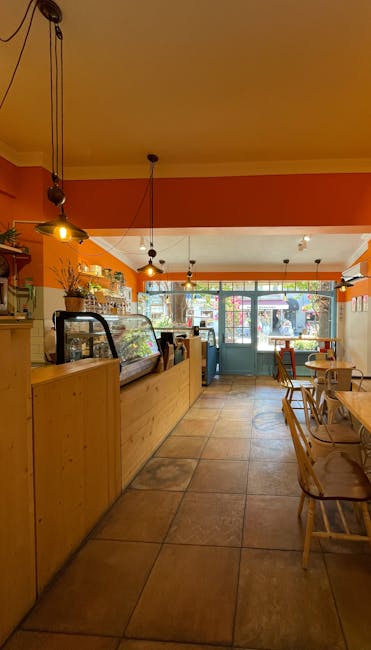 Inside a cafe with warm orange and yellow walls, pendant lights hanging from the ceiling, and large windows at the front allowing natural light to enter. The floor is tiled with a light brown pattern. A wooden counter with a glass display case containing food items is positioned on the left side, with various kitchen or storage items behind it. In the foreground, there are wooden chairs and tables arranged for seating. The scene is outside through the windows, showing a street view with parked cars and pedestrians, indicating that the cafe is located in a busy area. The image captures a clean, organized space in the process of either being prepared for a move or arranged for client service, with no visible boxes, packing materials, or moving equipment, suggesting it is a snapshot of the interior environment associated with local shop and café relocations handled by Man and a Van Kennington.