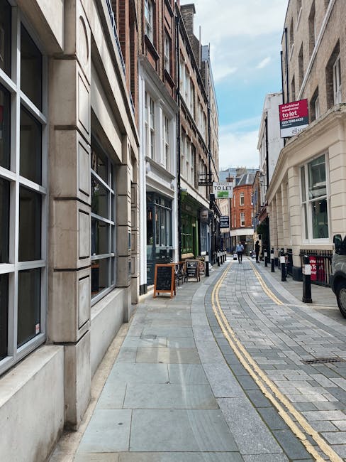 A narrow urban street scene showing the exterior façades of buildings on either side, with large glass windows and doors. The left side features a building with stone and concrete elements, while the right side includes a white-painted building with large windows. The pavement is paved with light and dark grey stone tiles, with double yellow lines running along the curb for parking restrictions. Several small black bollards line the right side of the street, and a few pedestrians are visible walking away in the distance. Overhead, there are signboards hanging from buildings advertising properties for sale or rent, including one visible which states 'TO LET'. The scene is well-lit with natural daylight, indicating a clear weather condition. The street appears to be in a commercial area, potentially suitable for house removals and furniture transport, as suggested by the context of property services provided by companies like Man and a Van Kennington.