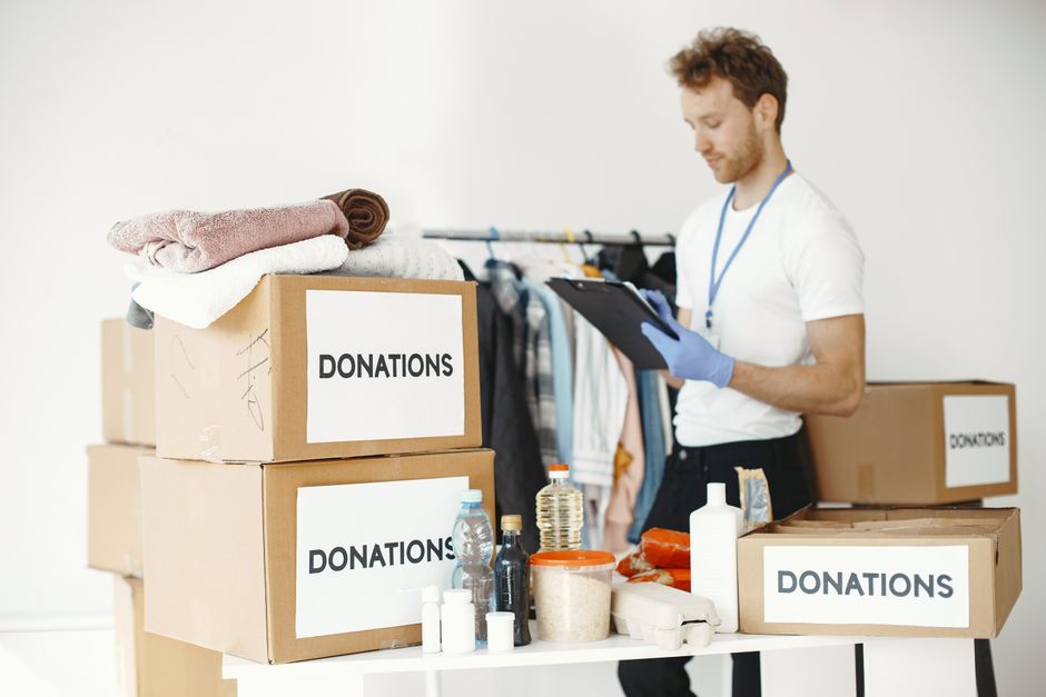 A young male volunteer wearing a blue t-shirt with 'VOLUNTEER' printed on it is participating in a house removal and waste disposal process. He is inside a property, holding a large black rubbish bag 