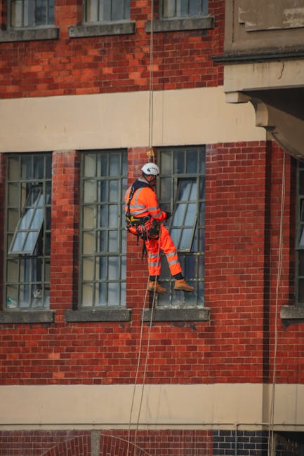 A worker in high-visibility orange safety gear, including a helmet and harness, is suspended by ropes in front of a red brick building during a house removal or building maintenance task. The worker appears to be descending or positioning near a large window with multiple panes, some of which are open, revealing an interior space. The background shows the façade of the building with brickwork and window casings, while the worker’s safety equipment, such as the harness and ropes, are clearly visible. This scene depicts a professional operation involving external access to a property, aligning with house removals or property maintenance services often provided by companies like Man and a Van Kennington. The natural lighting suggests daytime conditions, and the surrounding environment emphasizes the building's façade, windows, and the technician’s activity of working at height, likely as part of a relocation or external furniture transport process during home or business moving projects.
