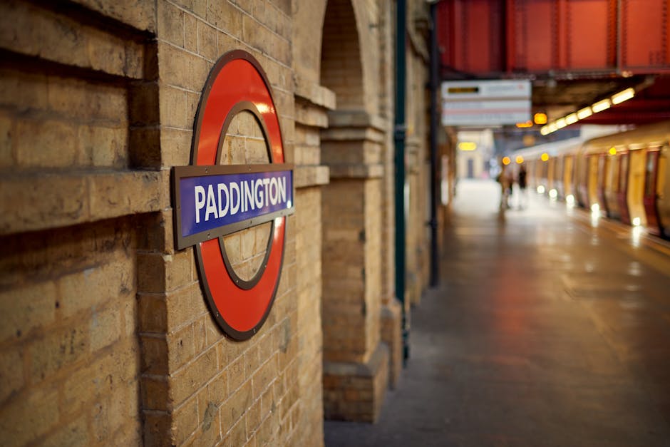 Inside Kennington Tube station platform, several passengers are waiting for their train, which displays the destination 'Kennington' on the overhead electronic sign, indicating an upcoming train arriv