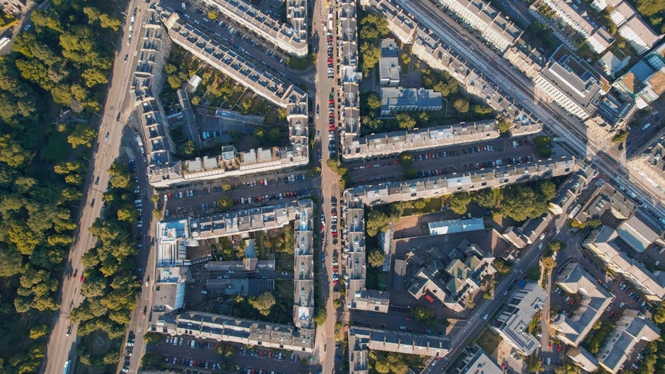 An aerial view of a residential area capturing rows of terraced houses with slate roofs, arranged in a grid-like pattern. The image shows a main road crossing through the neighborhood, lined with parked cars on both sides. To the left, there is a section of green trees providing foliage along the street, while on the right side of the image, larger commercial or office buildings are visible. The scene is well-lit with natural daylight, and the photograph illustrates the dense, urban environment typical of Kennington. This setting relates to home relocation and house removals, highlighting the urban context where [COMPANY_NAME] specializes in furniture transport, packing, and loading services for property moves such as the case study referenced in the 'Moving out of Kennington Oval flats removals' page.