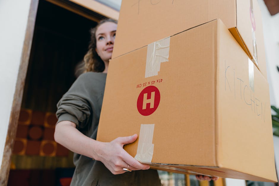A woman inside a property is holding a large cardboard box labeled with an 'H' and a red circle, part of a moving process. The woman, with curly hair and wearing a grey top, is positioned near a doorway or opening, with her arm extended to support the box, which is partially wrapped in packing tape. The box, marked with the word 'KITCHEN' written in blue marker, appears to contain household items, and she is in the act of carrying it, suggesting an organized home relocation and furniture transport scenario. Behind her, the interior features a wooden panel or ceiling, indicating she is inside a house or flat. Equipment such as trolleys, straps, or blankets are not visible, but the setting is consistent with professional removals, as provided by Man and a Van Kennington, offering packing and moving services. The lighting is natural, illuminating the scene for safe and efficient handling during the move.