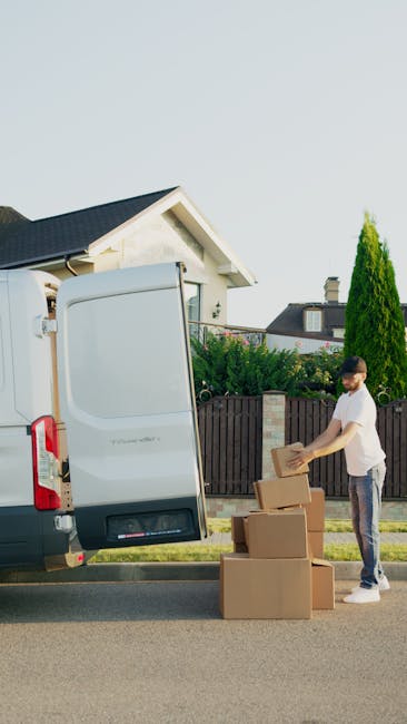 A man wearing a white t-shirt and jeans is standing on a residential street next to an open white van, which is parked on the pavement outside a house with a pitched roof and a chimney. The man is hol