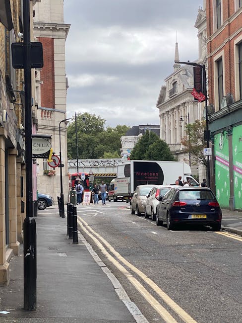 A narrow city street in Kennington during daytime, showing parked cars lining the curbside and a white moving truck from Man and a Van Kennington positioned to load or unload furniture and boxes. The street is bordered by historic red-brick and white buildings, with some featuring large windows and decorative architectural details. An orange and purple illuminated sign is mounted on a lamp post, and green and purple advertisements are visible on a building to the right. Pedestrians are walking near the truck, with some gathered around a doorway or in front of a shop. Overhead, a cloudy sky casts soft light over the scene. The area is marked by double yellow lines, indicating parking restrictions, supporting the need for careful planning in house relocation or furniture transport in tight urban spaces, and highlighting potential parking challenges often encountered during removals between residential and commercial properties in Kennington.