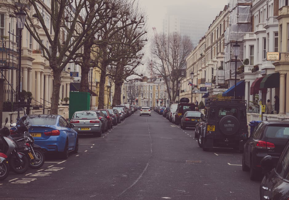 A narrow city street in Kennington during daytime, showing parked cars lining the curbside and a white moving truck from Man and a Van Kennington positioned to load or unload furniture and boxes. The street is bordered by historic red-brick and white buildings, with some featuring large windows and decorative architectural details. An orange and purple illuminated sign is mounted on a lamp post, and green and purple advertisements are visible on a building to the right. Pedestrians are walking near the truck, with some gathered around a doorway or in front of a shop. Overhead, a cloudy sky casts soft light over the scene. The area is marked by double yellow lines, indicating parking restrictions, supporting the need for careful planning in house relocation or furniture transport in tight urban spaces, and highlighting potential parking challenges often encountered during removals between residential and commercial properties in Kennington.