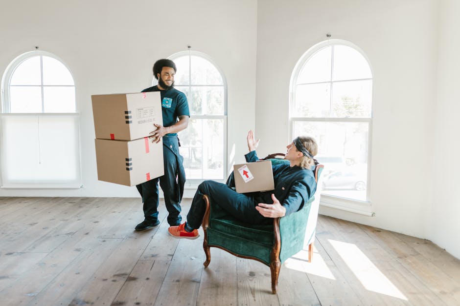 A man with a dark beard and dark hair, dressed in casual clothing, is inside a bright, minimalist room with white walls and large arched windows, carrying two medium-sized cardboard boxes sealed with red and black tape. He stands near an open doorway, smiling, as he prepares to load or move the boxes into the space. Meanwhile, a woman with short dark hair and glasses, wearing a dark jacket and red shoes, is seated in a vintage-style green armchair with wooden legs, holding a box with a white label and an upward-pointing arrow. She is raising her right hand in a stopping gesture while looking at the man, possibly indicating a pause or giving instructions during the packing and moving process. The room has light wooden flooring and natural daylight streaming in through the windows, reflecting the ongoing home relocation activities that Man and a Van Kennington provides as part of their removals service.